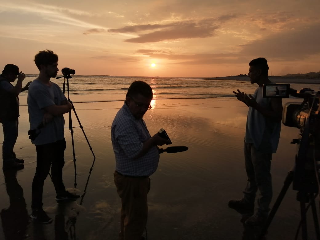 Man talking to people in front of sunset over water. John Kennedy at the estuarine border between Mexico and Guatemala filming an interview for his documentary Suchiate.
