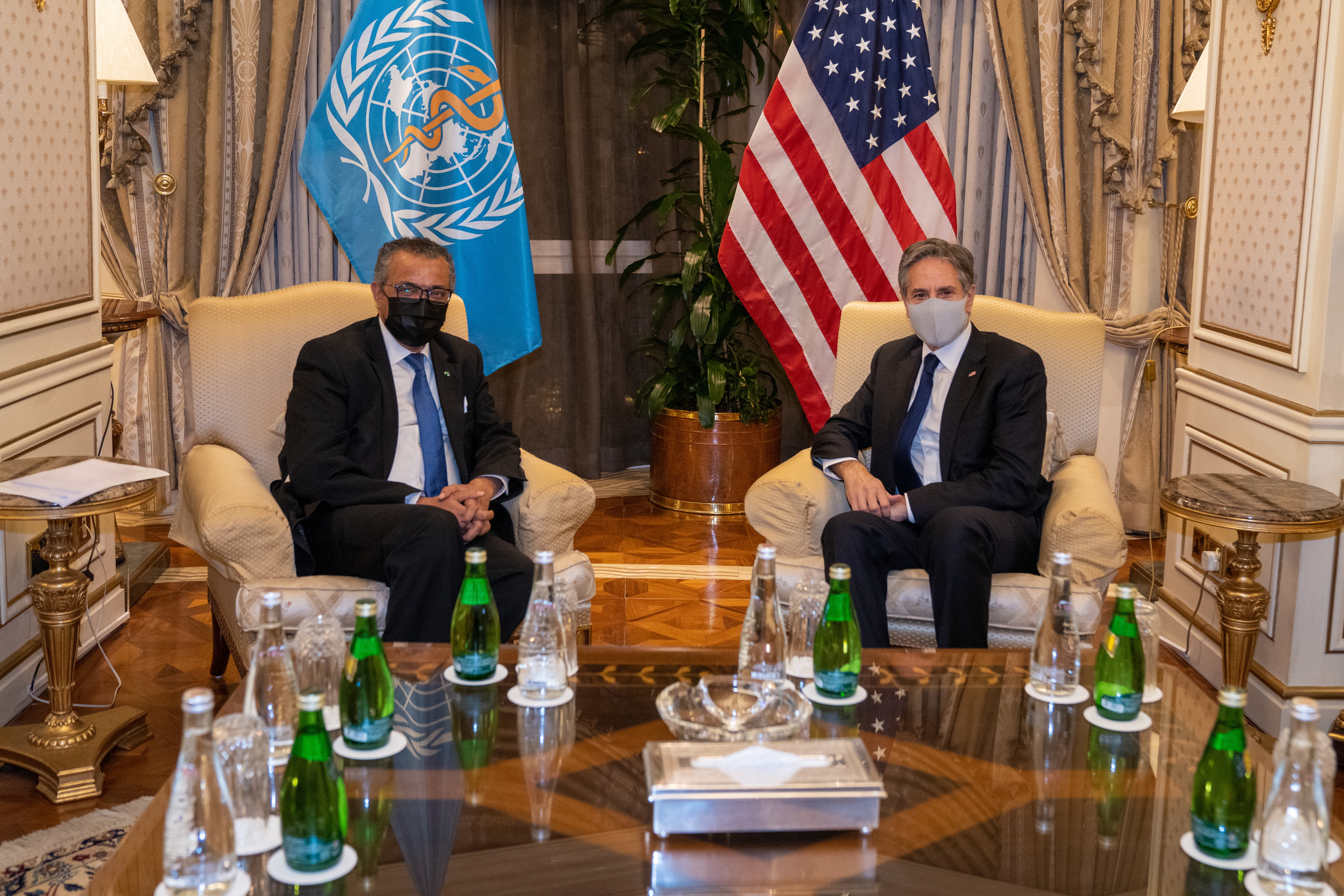 Secretary of State Antony J. Blinken meets with World Health Organization Director-General Dr. Tedros Adhanom Ghebreyesus wearing masks, seated in front of an American flag and a WHO flag. 