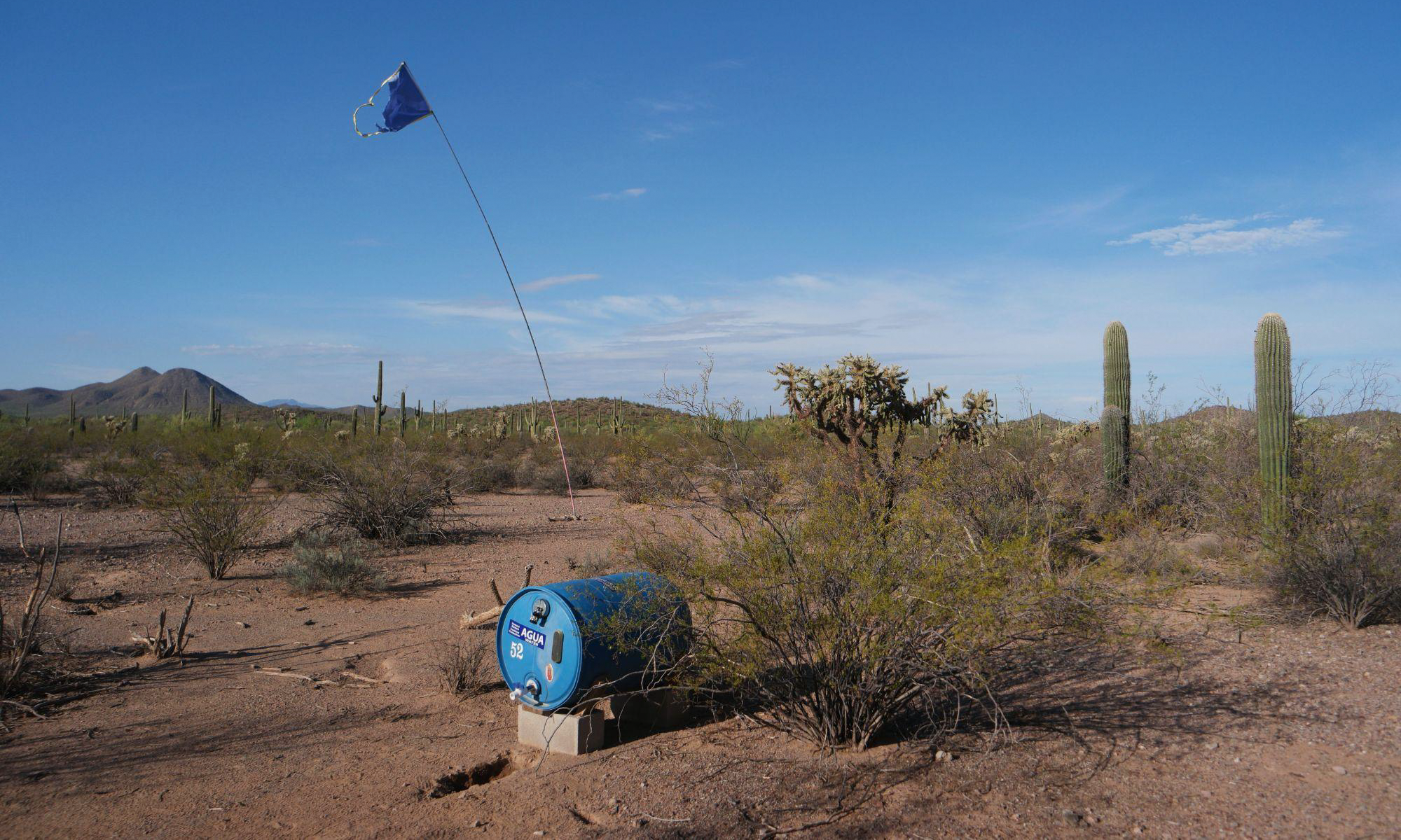 A blue water barrel with a tall flag sits in the middle of the desert. 