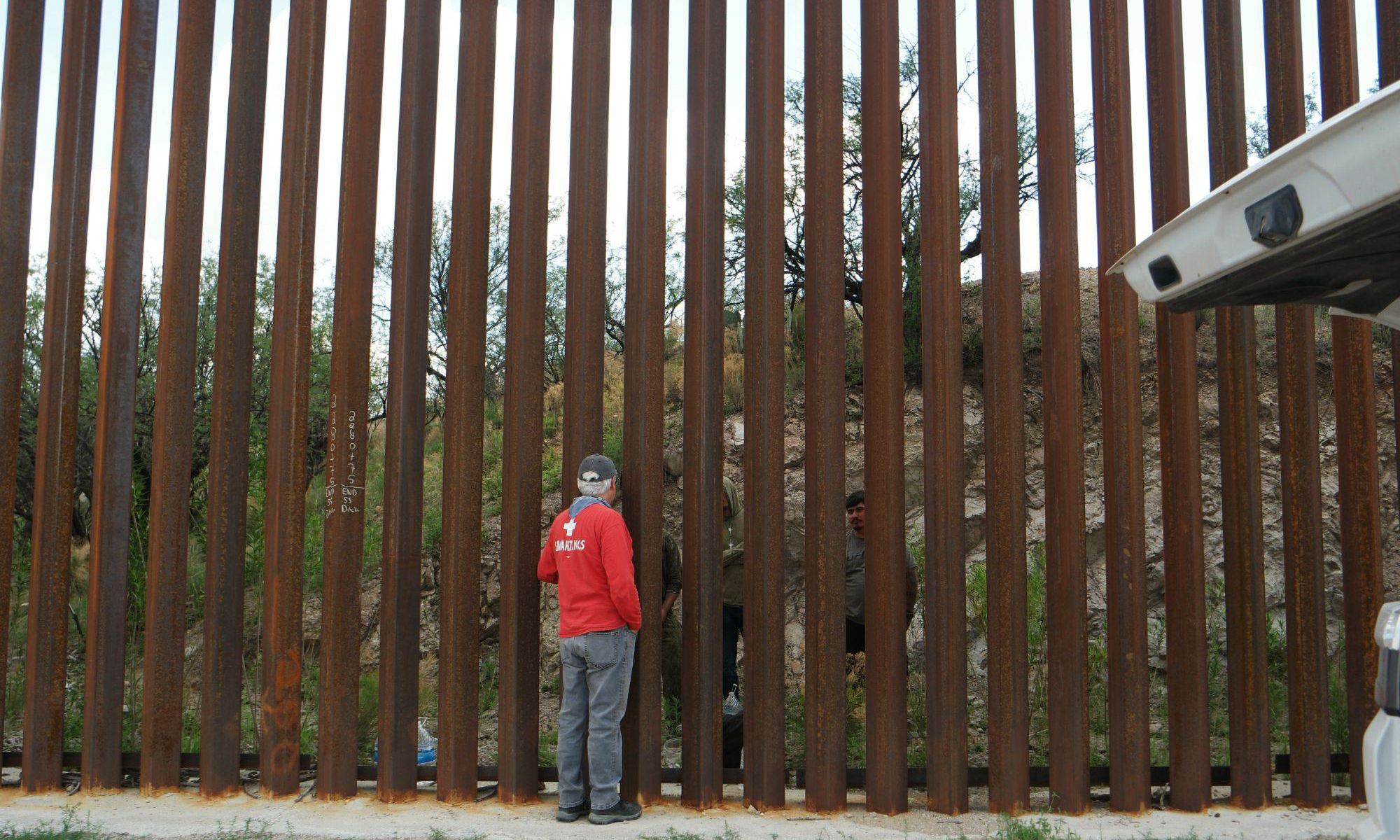 Men on both sides of a border wall speak through large iron posts.