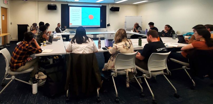 Scholar presents work in a slideshow, surrounded by a table of event attendees.