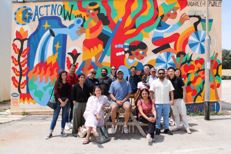 Spring school participants from Cornell and QMUL pose for a photo in front of a vibrant wall mural.