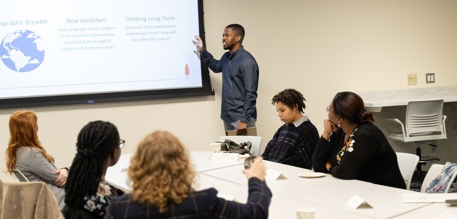Obioha Chijioke speaks to a small group while pointing toward a presentation slide.