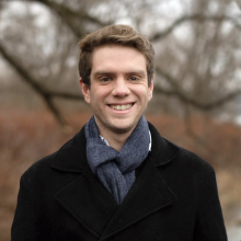 Casey with short cropped brown hair wears black jacket dark shirt smiles against a autumnal backdrop of trees and foliage